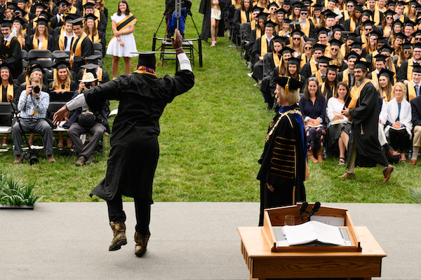 A student dancing while walking across the stage at commencement. He faces an audience of graduates and family members.