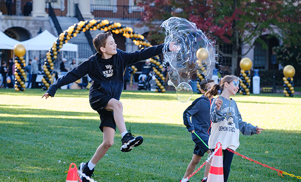 Children dressed in Wake Forest gold and black play with giant bubbles on the Quad. Behind are gold and black balloons